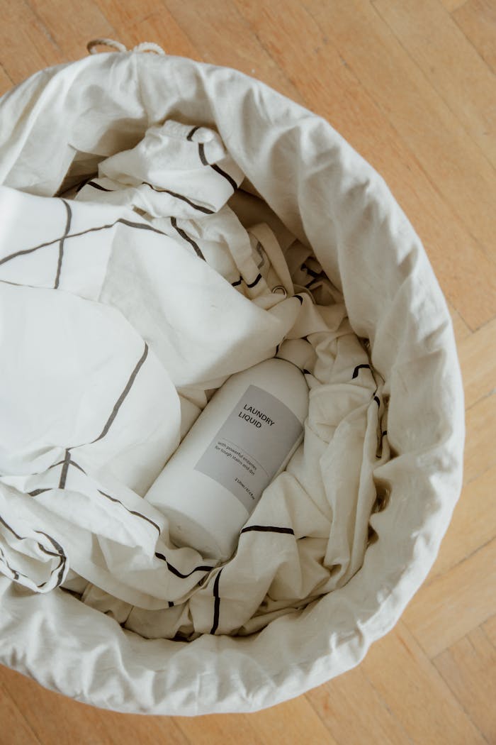 Overhead view of a laundry basket filled with cloths and a detergent bottle on wooden floor.